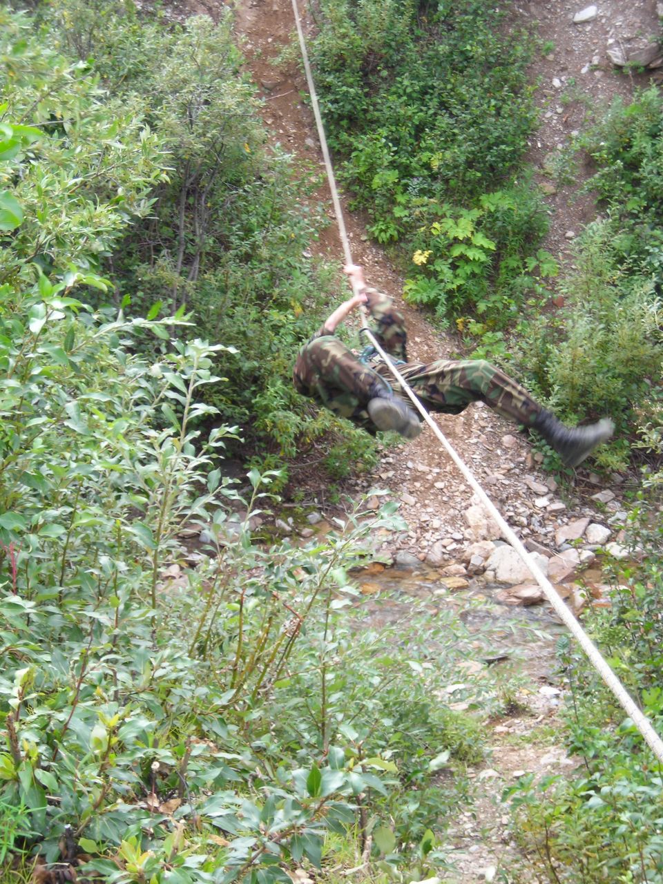 Person in camouflage rappelling down a rocky, wooded slope with a rope
