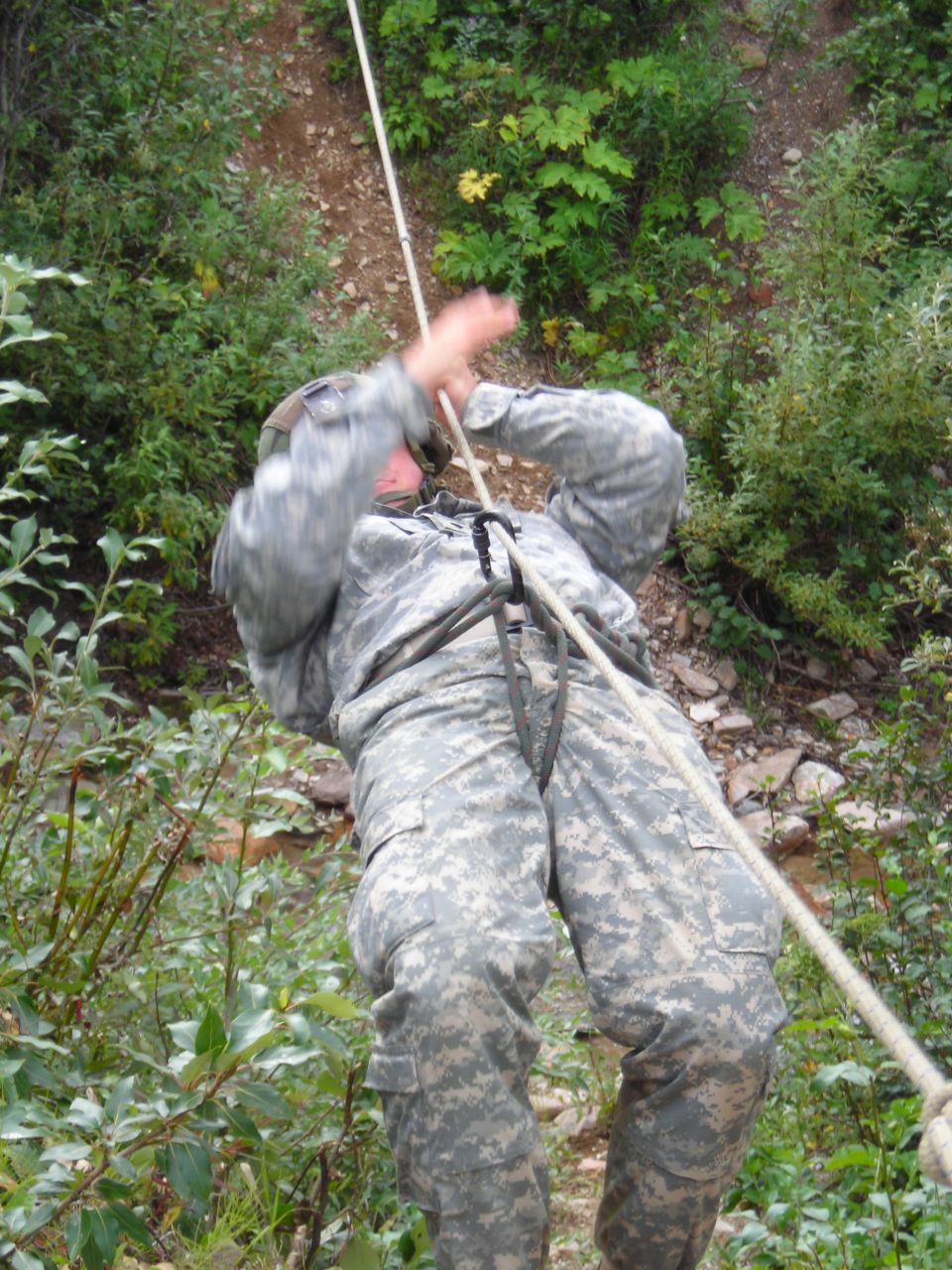 Soldier in camouflage uniform rappelling down a rocky, wooded slope using a rope