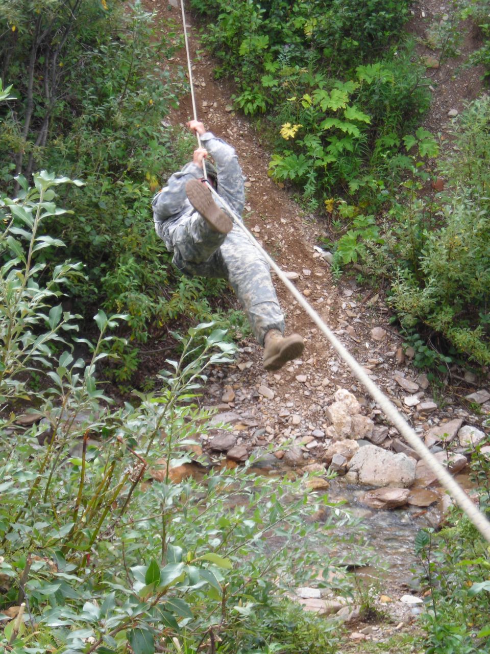 Soldier in camouflage rappelling down a rocky, wooded slope using a rope. A small stream is visible at the bottom