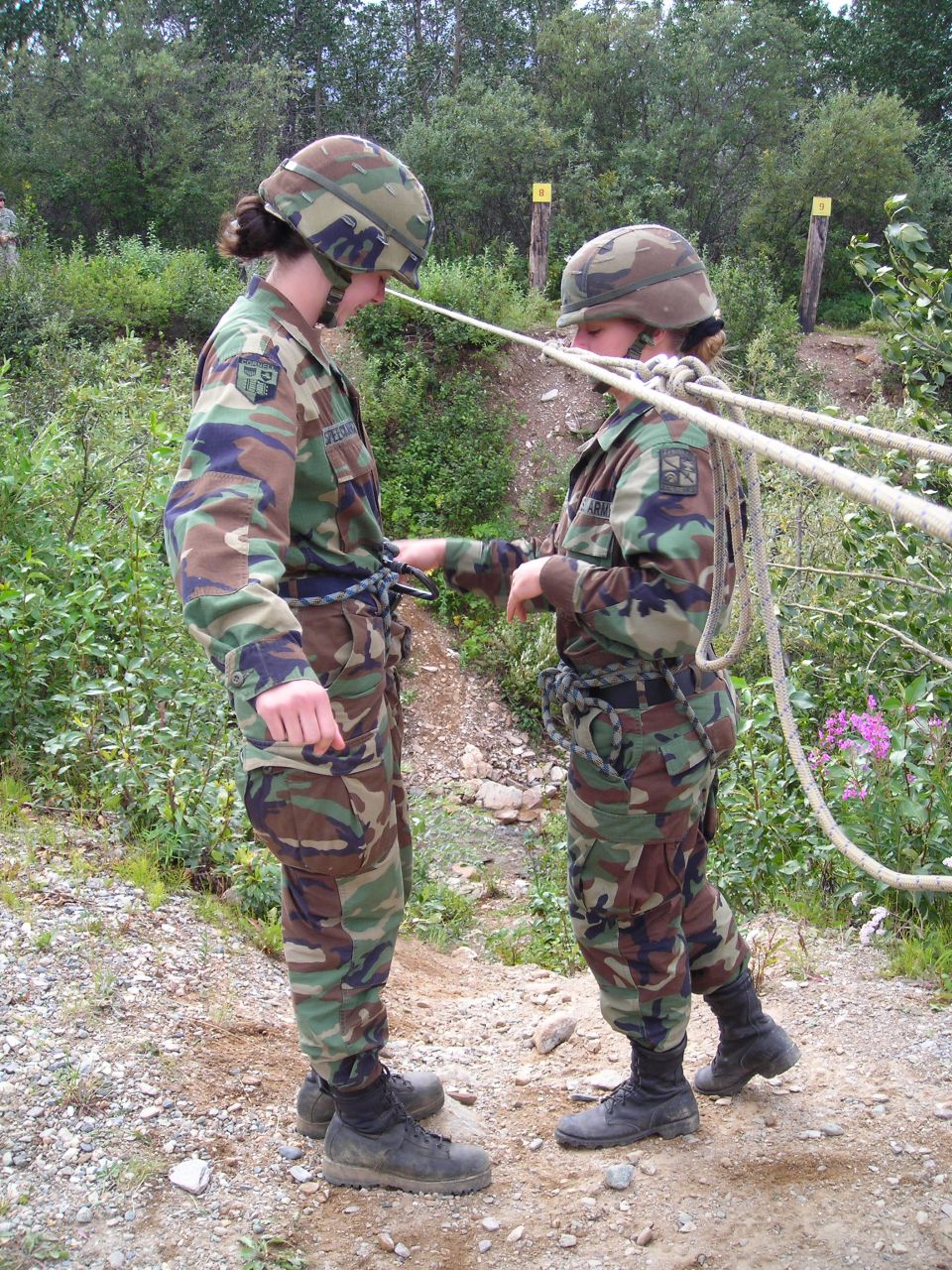 Two soldiers in camouflage uniforms and helmets practice with ropes outdoors on a dirt path