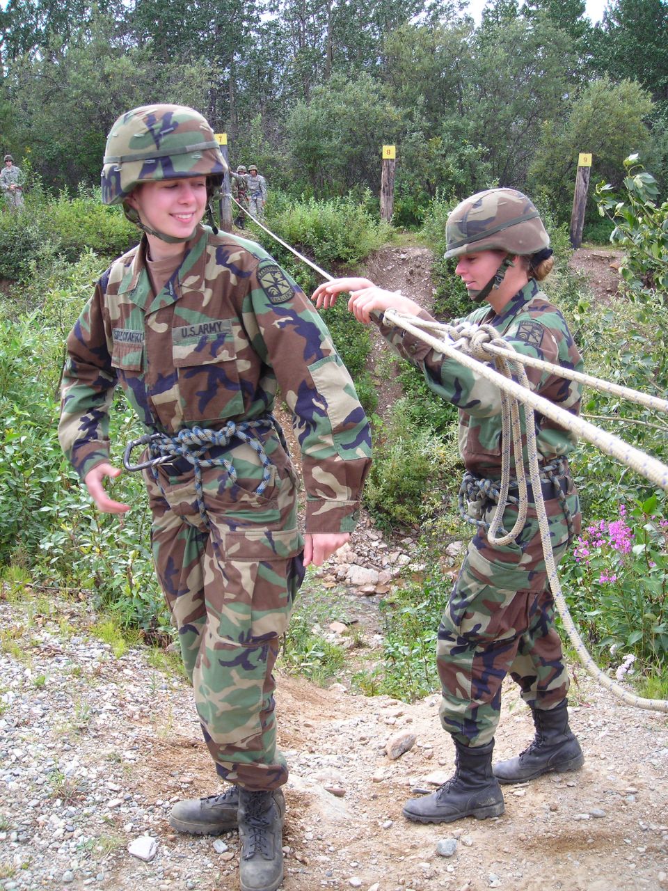 Two soldiers in camouflage uniforms and helmets practice with ropes outdoors. One soldier is in the foreground, smiling, with ropes tied around their waist. The other soldier is behind and to the right, holding the ropes