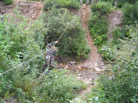 2003 Jeep TJ - A person in camouflage gear traverses a rocky stream bed using a rope for support.