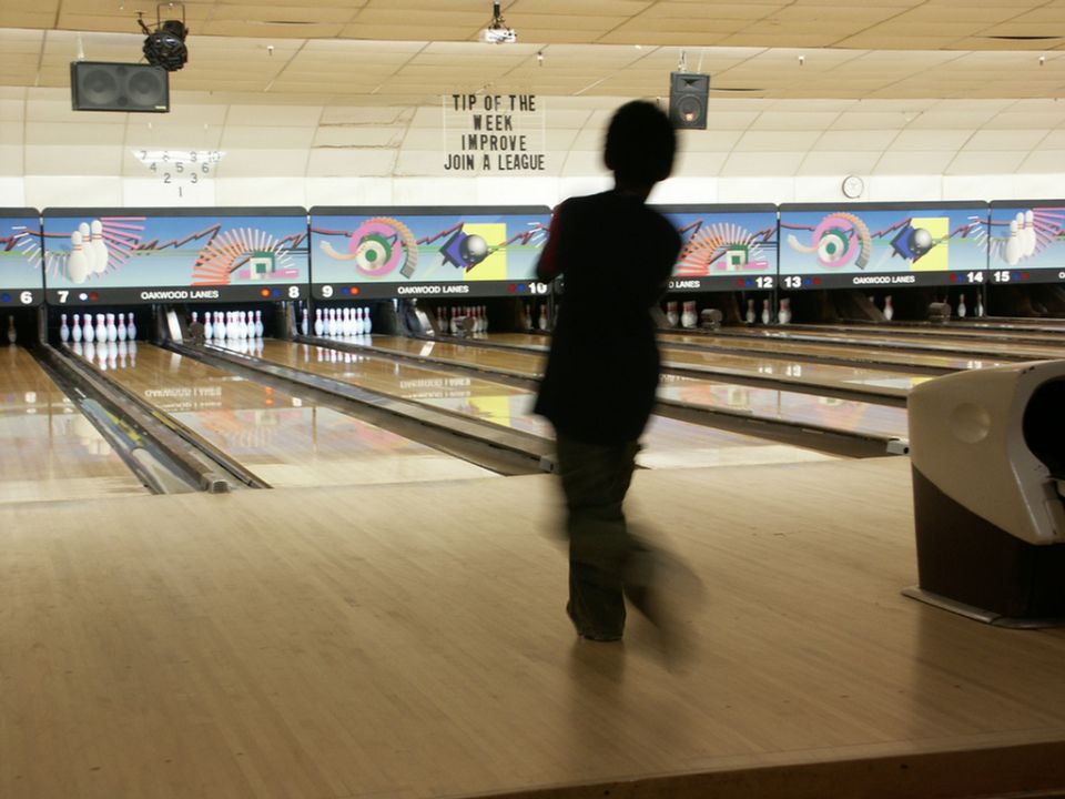 Bowling alley interior with lanes, pins, and a person bowling. Text on wall reads 'TIP OF THE WEEK IMPROVE JOIN A LEAGUE'