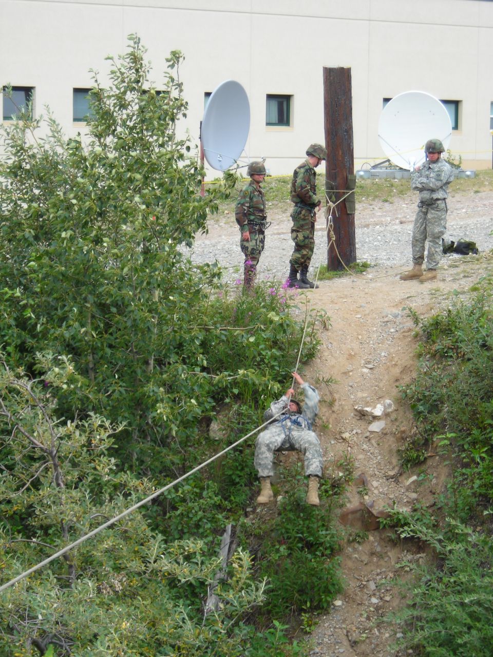 Soldiers practice rappelling down a dirt slope using a rope, with others observing and a building with satellite dishes in the background