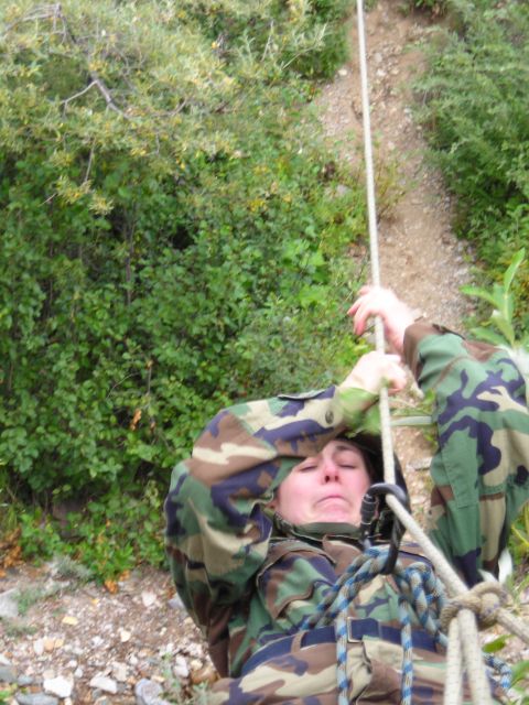 2003 Jeep TJ - A person in camouflage gear is rappelling down a steep, rocky incline with trees and brush surrounding the area.