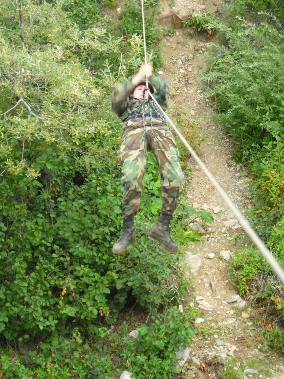 Person in camouflage rappelling down a rocky, vegetated slope using a rope