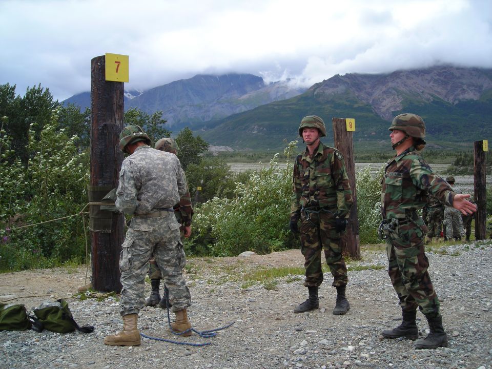 Soldiers in camouflage uniforms stand near wooden posts with yellow markers numbered 7 and 8 during an outdoor training exercise