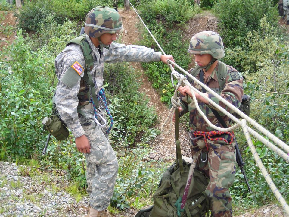 Two soldiers in camouflage uniforms and helmets practice rappelling with ropes and gear on a wooded hillside