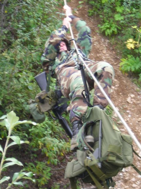 2003 Jeep TJ - A soldier in camouflage gear descends a steep, wooded incline using a rope, equipped with a rifle and a large backpack.