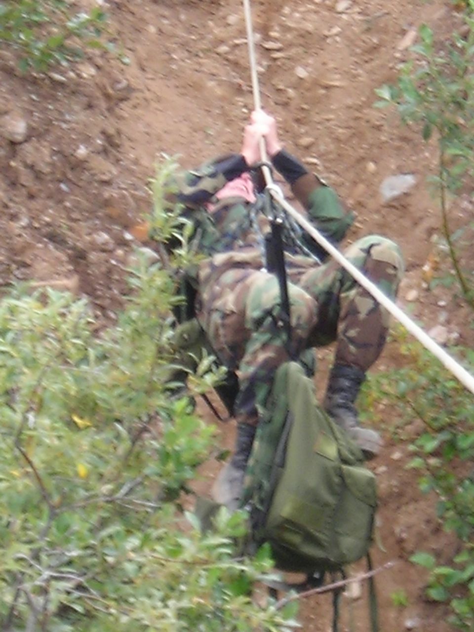 Person in camouflage rappelling down a dirt embankment with a rope and backpack