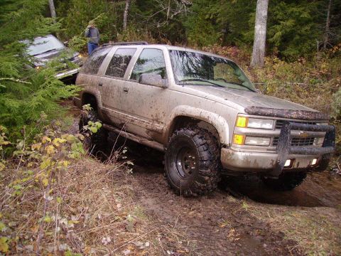 2000 Chevrolet Tahoe - A light-colored SUV is stuck in the mud on a wooded trail, with another vehicle partially visible behind it.