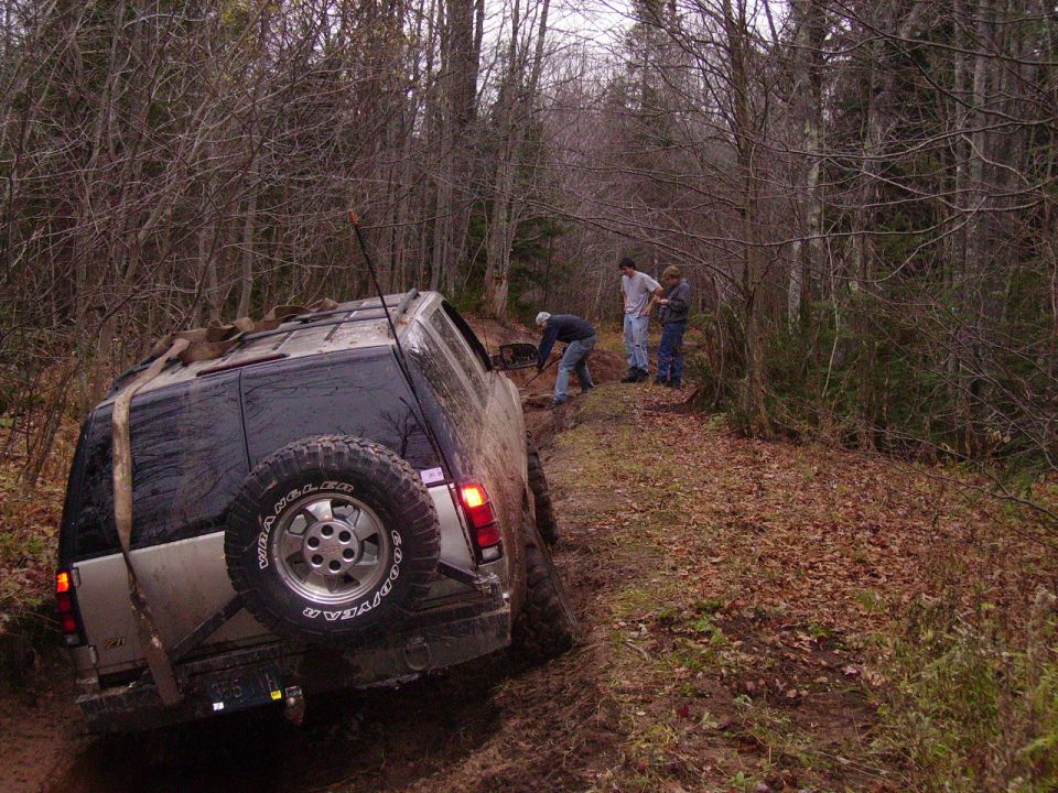SUV stuck in mud on a wooded trail, people trying to dig it out