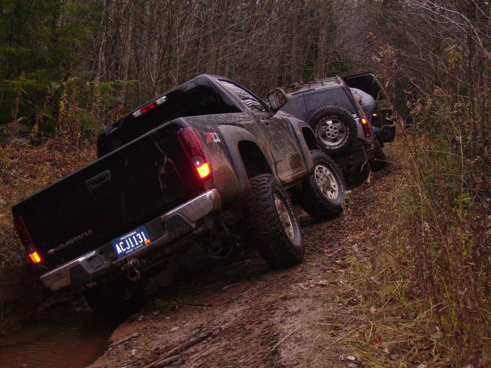 Muddy black pickup truck and SUV tilted on a dirt path with trees in the background