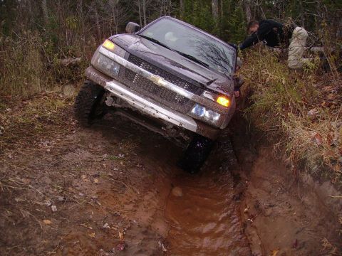 2000 Chevrolet Tahoe - A dark SUV navigates a challenging muddy terrain, with one person attempting to assist.