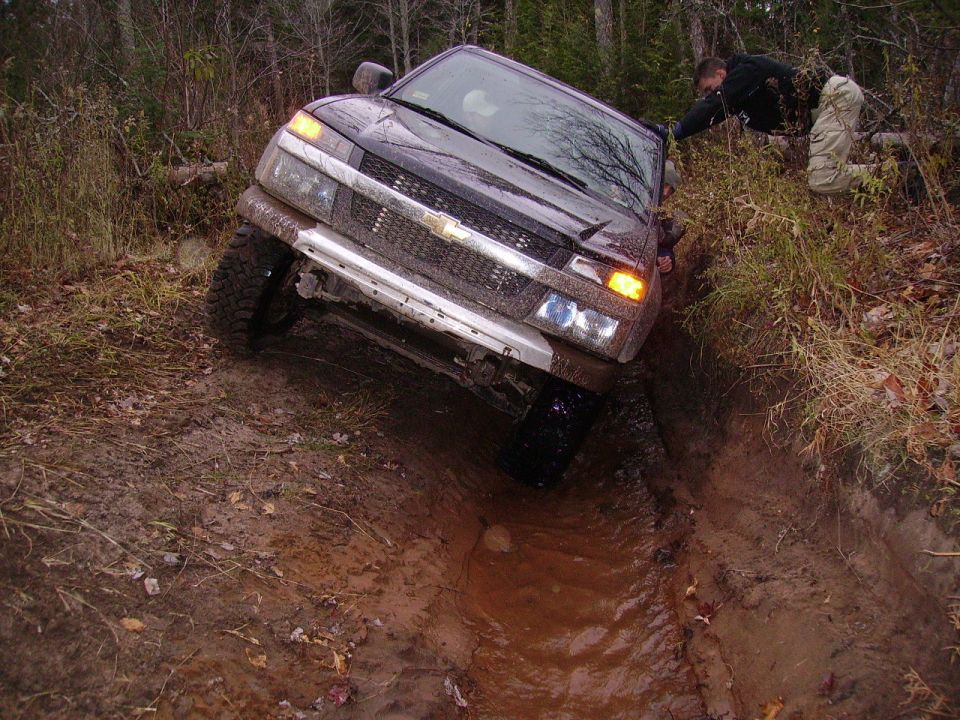 Dark SUV with a silver grille stuck in a muddy trench, front-left tire submerged, a person reaching towards it