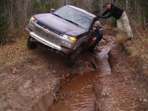 2000 Chevrolet Tahoe - A dark pickup truck is stuck in a muddy rut with two people attempting to push it out.