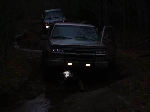 2000 Chevrolet Tahoe - Two SUVs navigate a muddy trail in the dark, with one vehicle's door ajar.