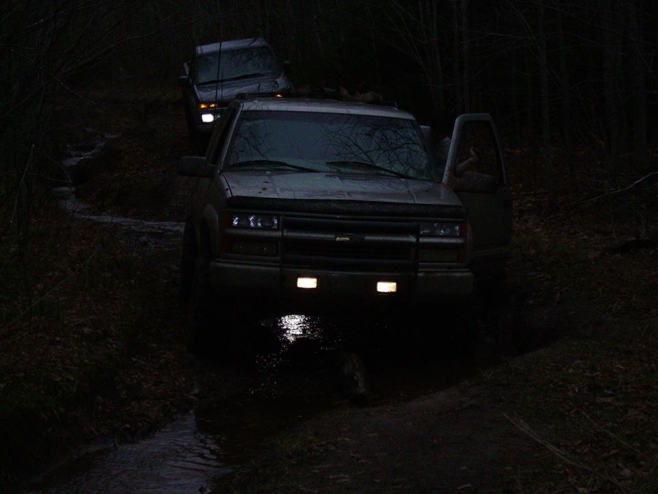 two SUVs driving through mud and water at night, one with the driver's door open