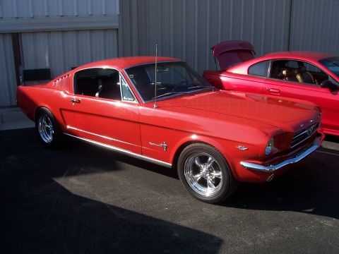 2002 Ford Mustang - A classic red Ford Mustang is parked next to a newer model Mustang. The older car has a chrome bumper and classic wheels.