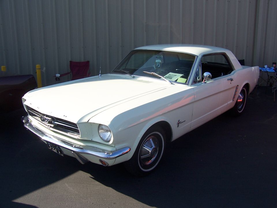 White early Ford Mustang hardtop coupe at a car show beside a metal building