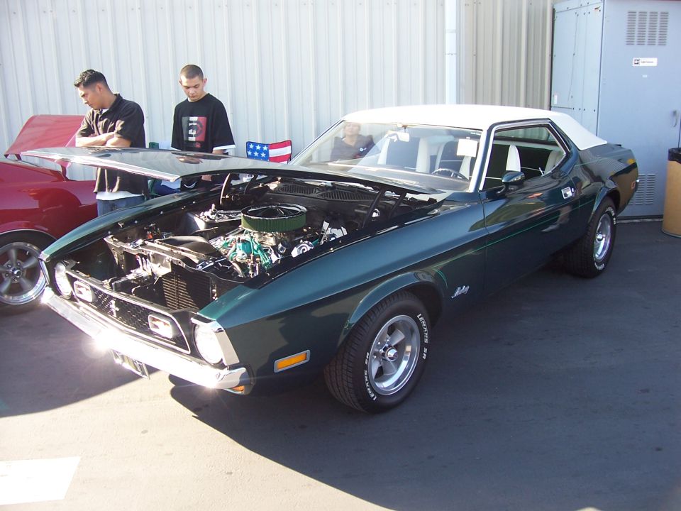 Green Mustang with white vinyl top, hood open showing engine. Two guys looking at it. Red car to the left