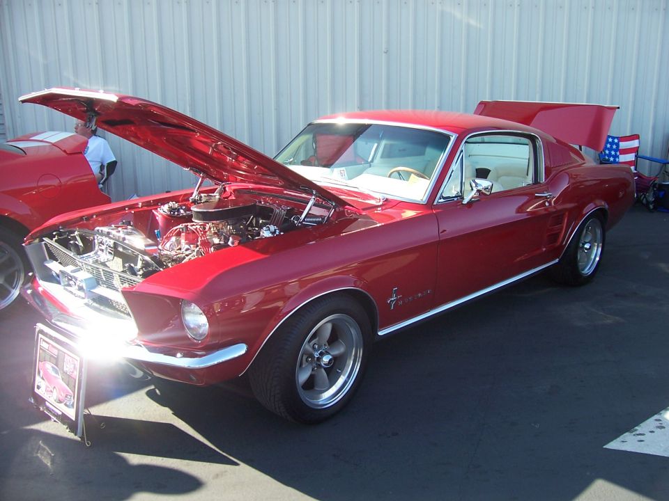 Red 1960s Mustang fastback, hood open over V8, on Torq Thrust wheels at a show