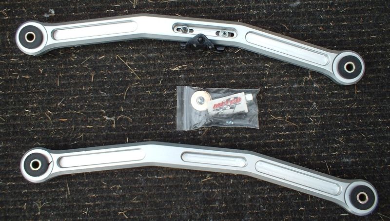 Two aluminum aftermarket suspension control arms with bushings and a bag of hardware on carpet