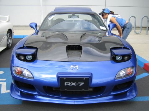 1993 Mazda RX-7 - A blue Mazda RX-7 is parked in a lot, with a woman leaning over to look at the engine bay.