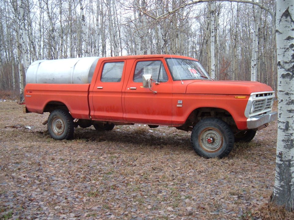 Orange crew cab pickup truck with a large silver tank in the bed, parked in a wooded area