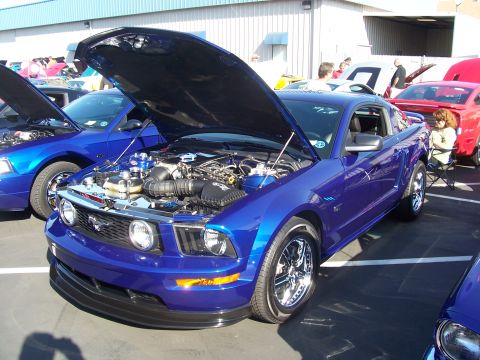 2002 Ford Mustang - A blue Ford Mustang is displayed with its hood open, revealing the engine compartment. Other vehicles are parked nearby.