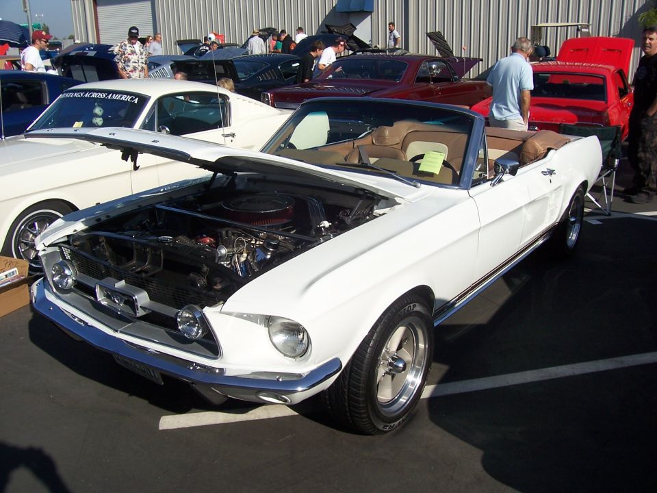White 1967-1968 Ford Mustang convertible with hood open, showing engine. Other cars are parked nearby