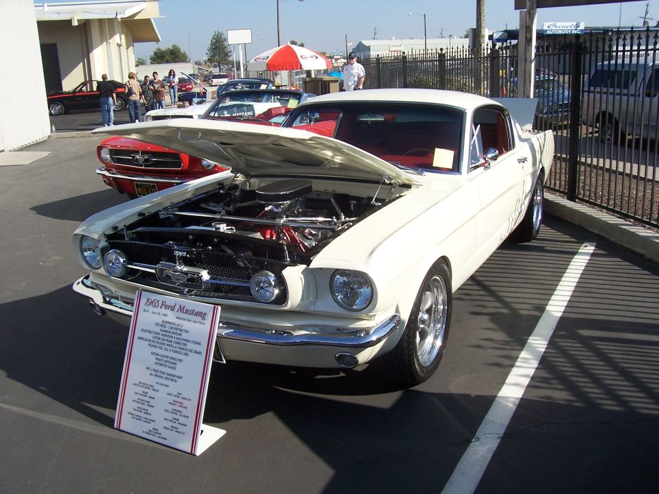 White 1965 Ford Mustang with hood open, parked next to a red Mustang. Sign in front details the car