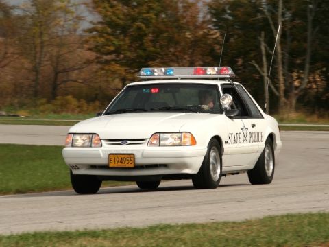 1993 Ford Mustang - A white 1993 Ford Mustang police car with a light bar on the roof drives down a road.