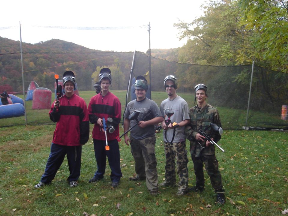 Five young men in paintball gear posing on a grassy field with autumn foliage