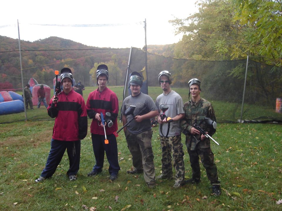 Five men in paintball gear stand in a grassy field with trees and a net in the background