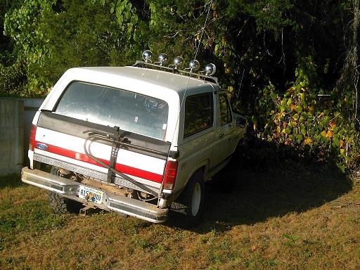White Ford Bronco, rear view, parked on grass with trees behind. Red stripe on tailgate