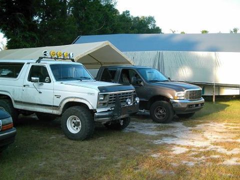 1986 Ford Bronco - Two Ford trucks, a Bronco and an F-250, are parked in front of large covered structures.