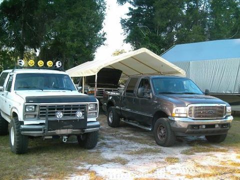 1986 Ford Bronco - Two trucks are parked in a lot, one is a white SUV and the other is a dark gray pickup truck.