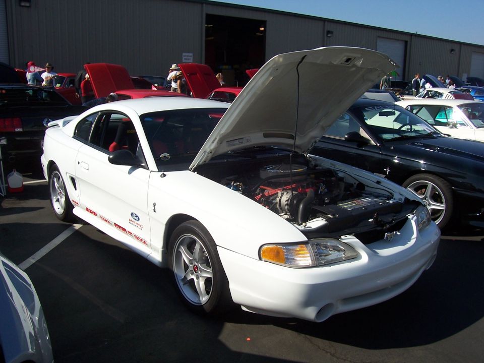 White Ford Mustang with hood open, showing engine. Red interior visible. Side graphics read '4.6L SUPERCHARGED'. Black Mustang parked behind
