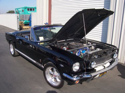 2002 Ford Mustang - A black 1965 Ford Mustang convertible is parked outside a garage with its hood open, revealing the engine bay.