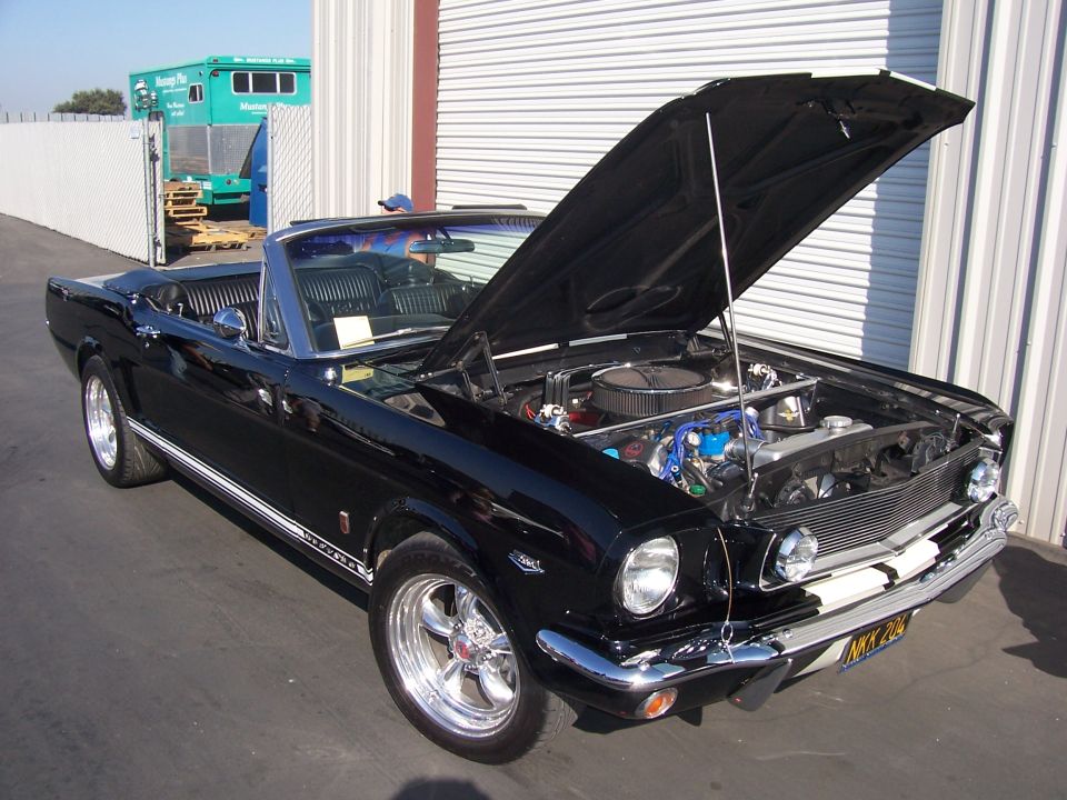 Black 1960s Mustang convertible with hood open showing V8 engine, parked outdoors