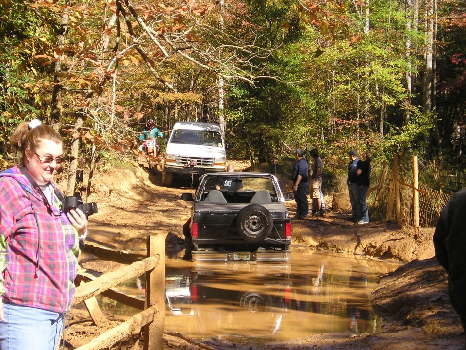 1996 Ford Bronco driving through mud, with a white van behind it on a wooded trail