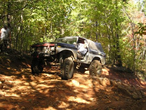 1996 Ford Bronco - A modified SUV navigates a challenging off-road trail surrounded by trees.