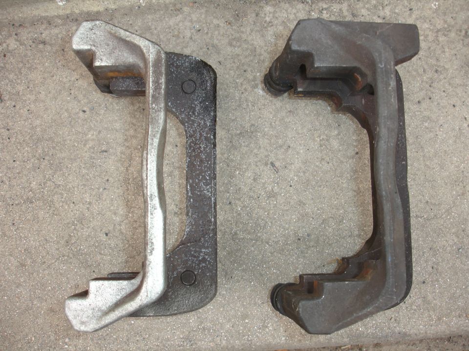 Two metal brake caliper brackets, one silver and one dark gray, sit on a concrete surface