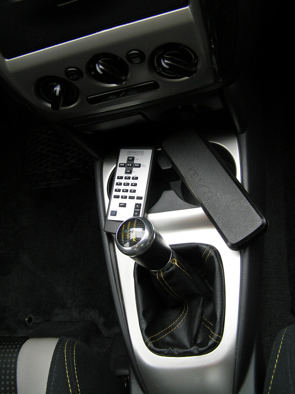 Car interior shot showing a manual gear shift, Kenwood remote, and cup holder