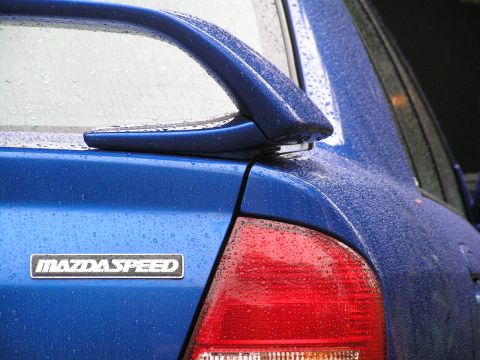 2003 Mazda Protege - A blue MazdaSpeed sedan's rear is visible, with raindrops on the paint and a prominent spoiler.