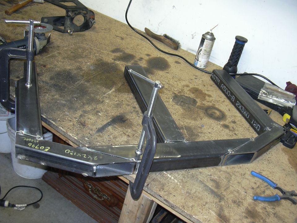 Metal fabrication project clamped to workbench, with tools and spray can nearby. Text on metal reads '2X2X120' and 'ASTM A500-B'