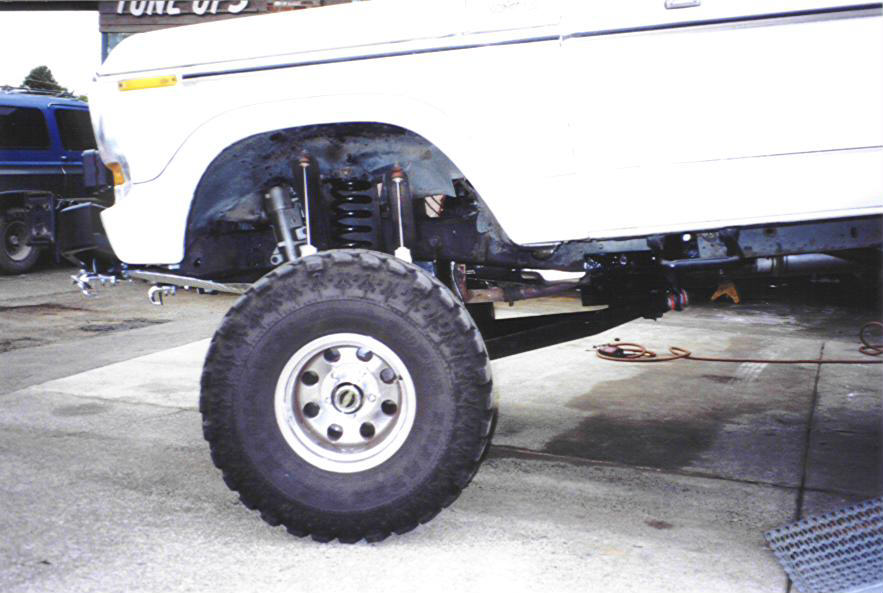 White SUV's front wheel and suspension system are visible, with a blue SUV in the background