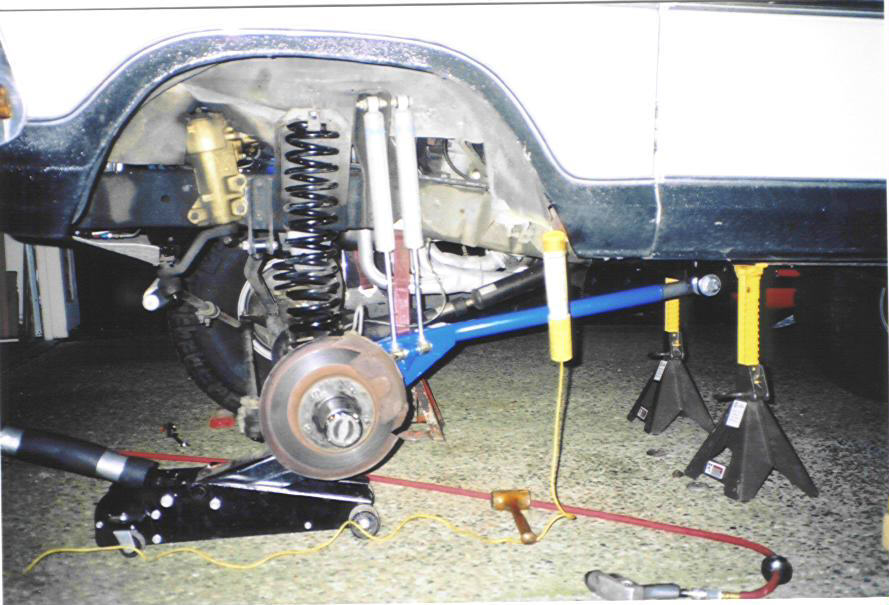 Vehicle's rear suspension is disassembled, with shocks, springs, and control arms visible. A floor jack and jack stands support the vehicle