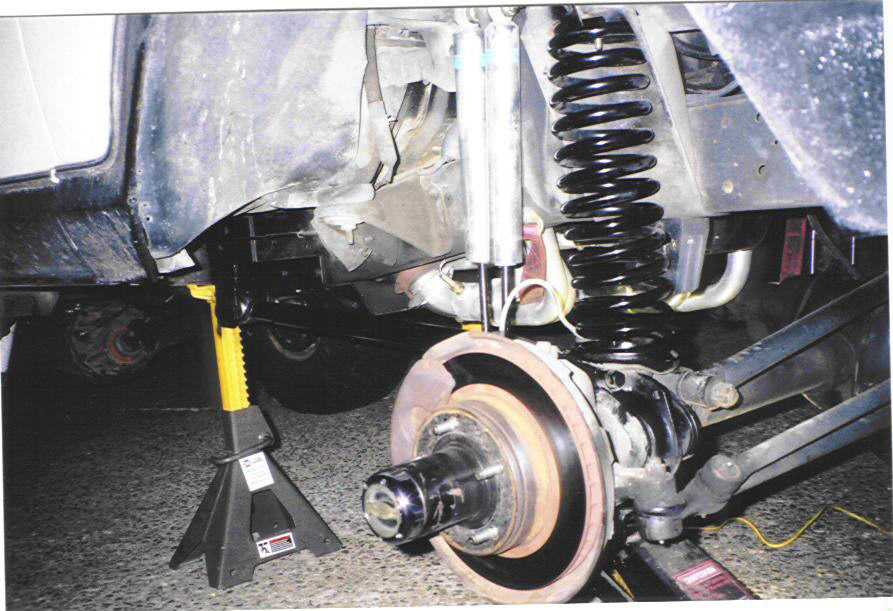 Close-up of a vehicle's front suspension with a jack stand supporting the frame. Black coil spring and silver shock absorber are visible